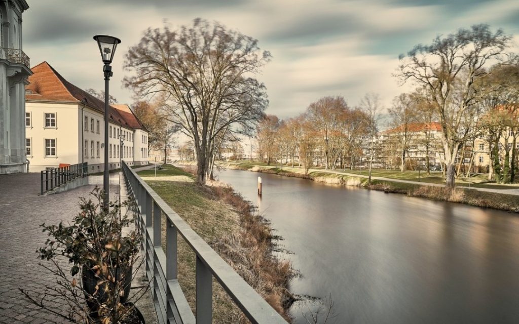 Spokojny widok na rzekę Hawelę w Oranienburgu, z nadrzeczną promenadą, zabytkową zabudową, nagimi drzewami i miękko rozmytą taflą wody