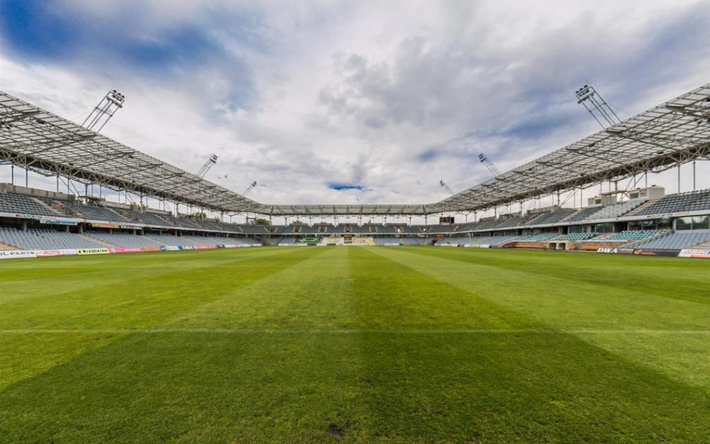 Stadion piłkarski z zieloną murawą, zadaszonymi trybunami oraz pustymi miejscami dla kibiców pod zachmurzonym niebem