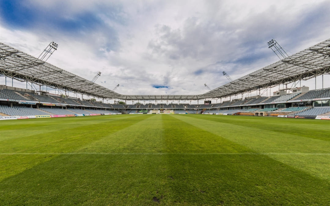 Stadion piłkarski z zieloną murawą, zadaszonymi trybunami oraz pustymi miejscami dla kibiców pod zachmurzonym niebem