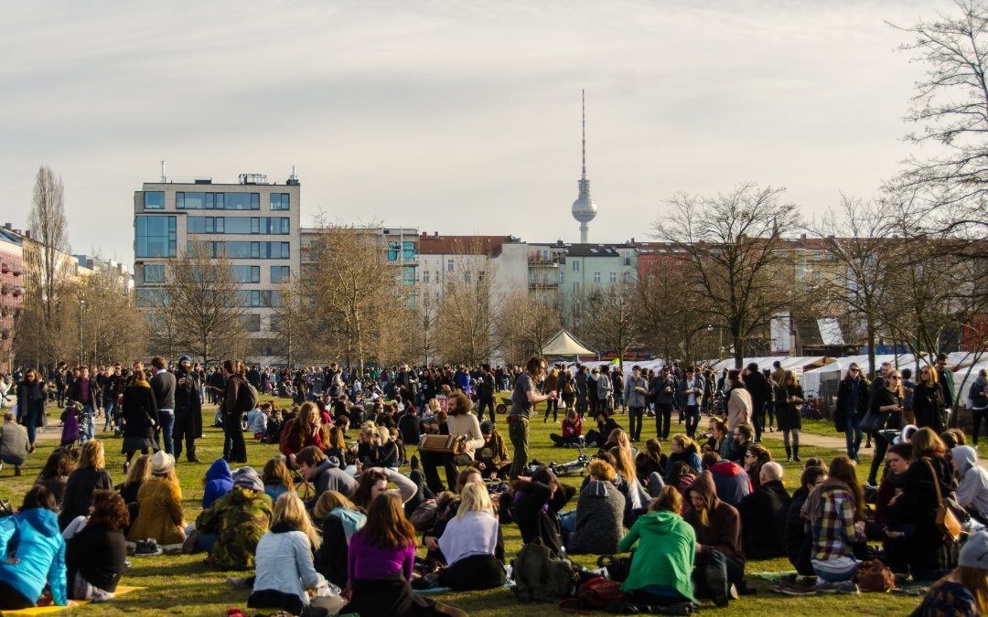 Tłum odpoczywających ludzi w Mauerpark w Berlinie, z zieloną łąką, stoiskami targowymi i wieżą Fernsehturm widoczną w oddali