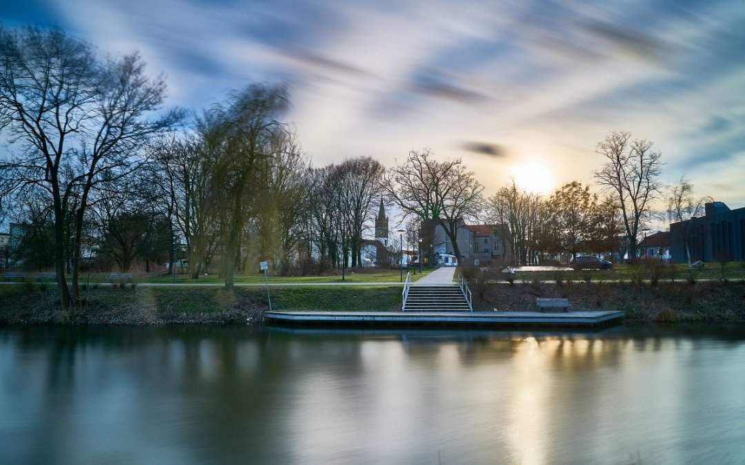 Widok na rzekę Hawelę w Oranienburgu o zachodzie słońca, z przystanią, schodami prowadzącymi nad wodę, drzewami i kościelną wieżą w tle
