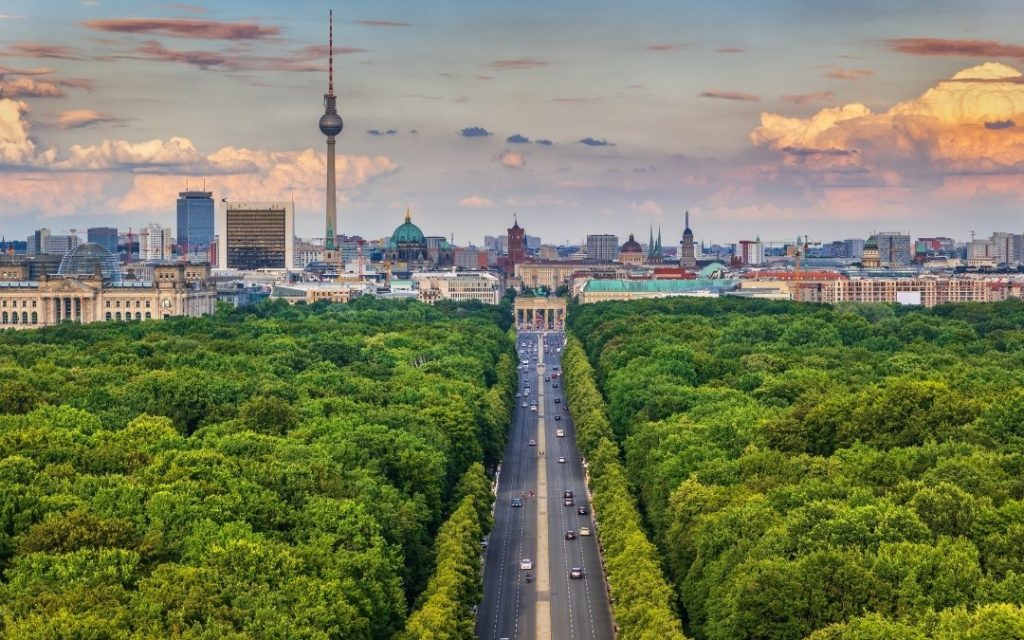 Widok z góry na Berlin ponad zielenią Tiergarten, z aleją prowadzącą do Bramy Brandenburskiej, wieżą Fernsehturm i zachmurzonym niebem nad centrum miasta