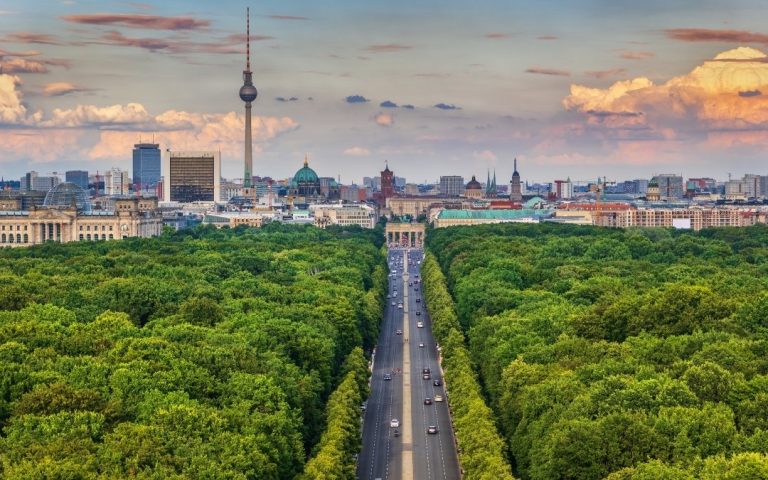 Widok z góry na Berlin ponad zielenią Tiergarten, z aleją prowadzącą do Bramy Brandenburskiej, wieżą Fernsehturm i zachmurzonym niebem nad centrum miasta