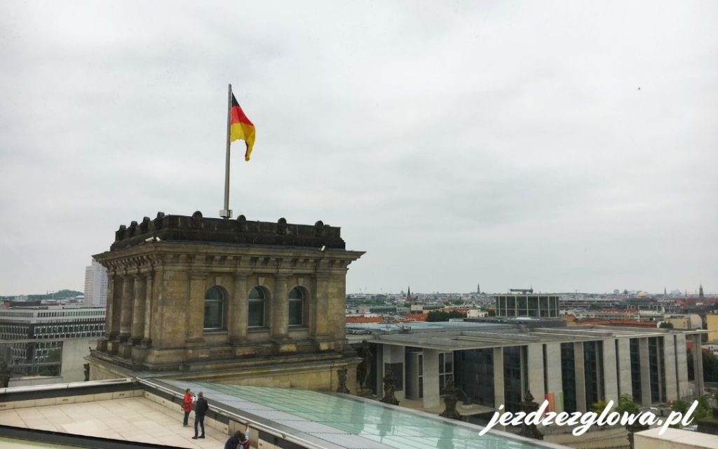 Widok z tarasu Reichstagu w Berlinie na zabytkową część budynku z niemiecką flagą oraz panoramę dzielnicy