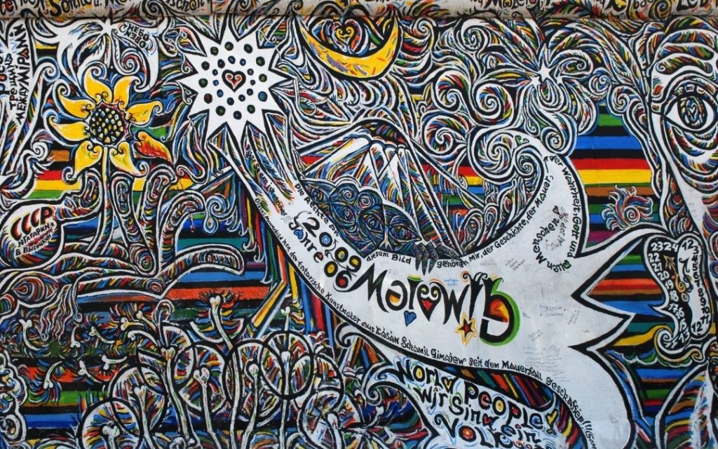 Zbliżenie na mural przy East Side Gallery w Berlinie, z kolorowymi spiralnymi wzorami, słonecznikiem, białym ptakiem i napisami pokrywającymi fragment Muru Berlińskiego