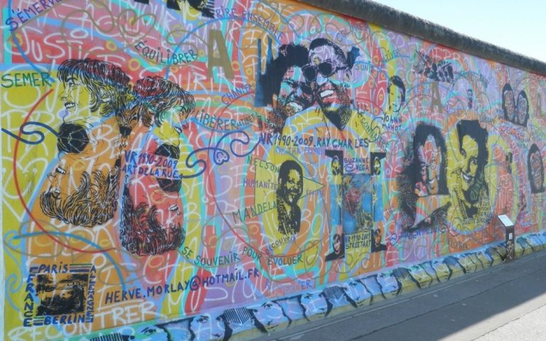 mural na Murze Berlińskim przy East Side Gallery, z portretami postaci, napisami i wielobarwnym tłem pełnym linii oraz graffiti