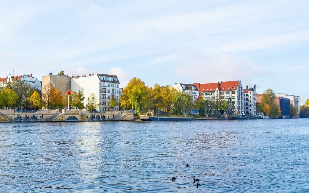 widok na nabrzeże Sprewy w Berlinie, z kamienicami, jesiennymi drzewami i kaczkami pływającymi po wodzie